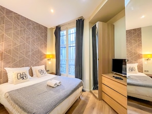 Una habitación de hotel con cama, televisión, cómoda de madera y ventana con cortinas.