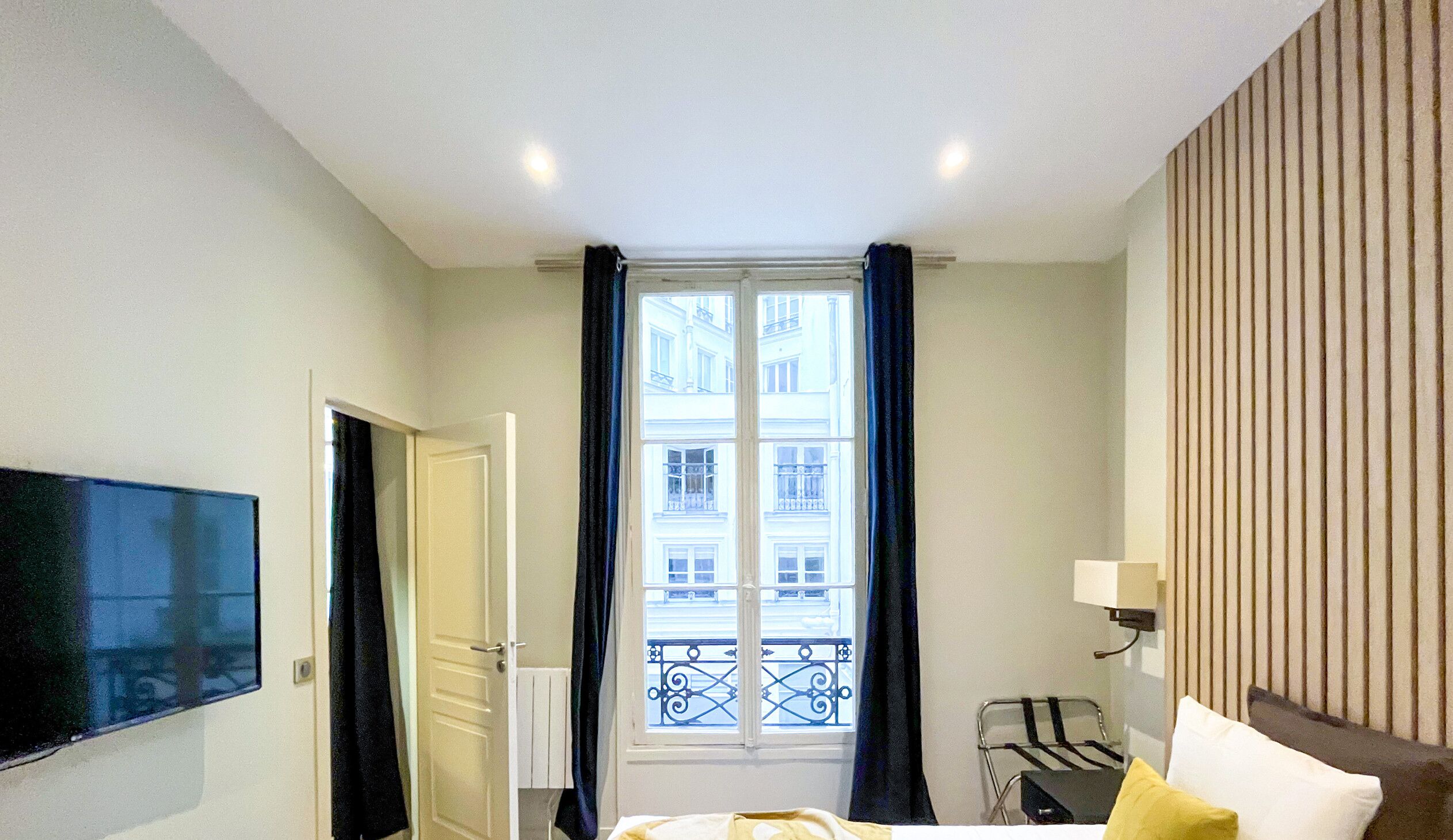 Une chambre d’hôtel avec un lit, un téléviseur fixé au mur, une fenêtre avec des rideaux, un radiateur et une petite table avec une chaise.