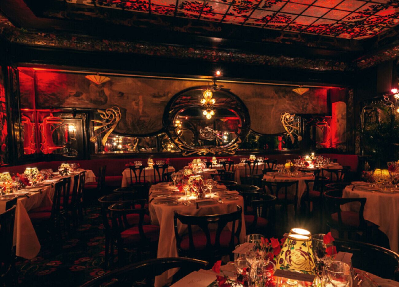 Un restaurant à la lumière tamisée, avec des tables rondes dressées pour le dîner, agrémenté de décorations murales élaborées et d’un lustre.