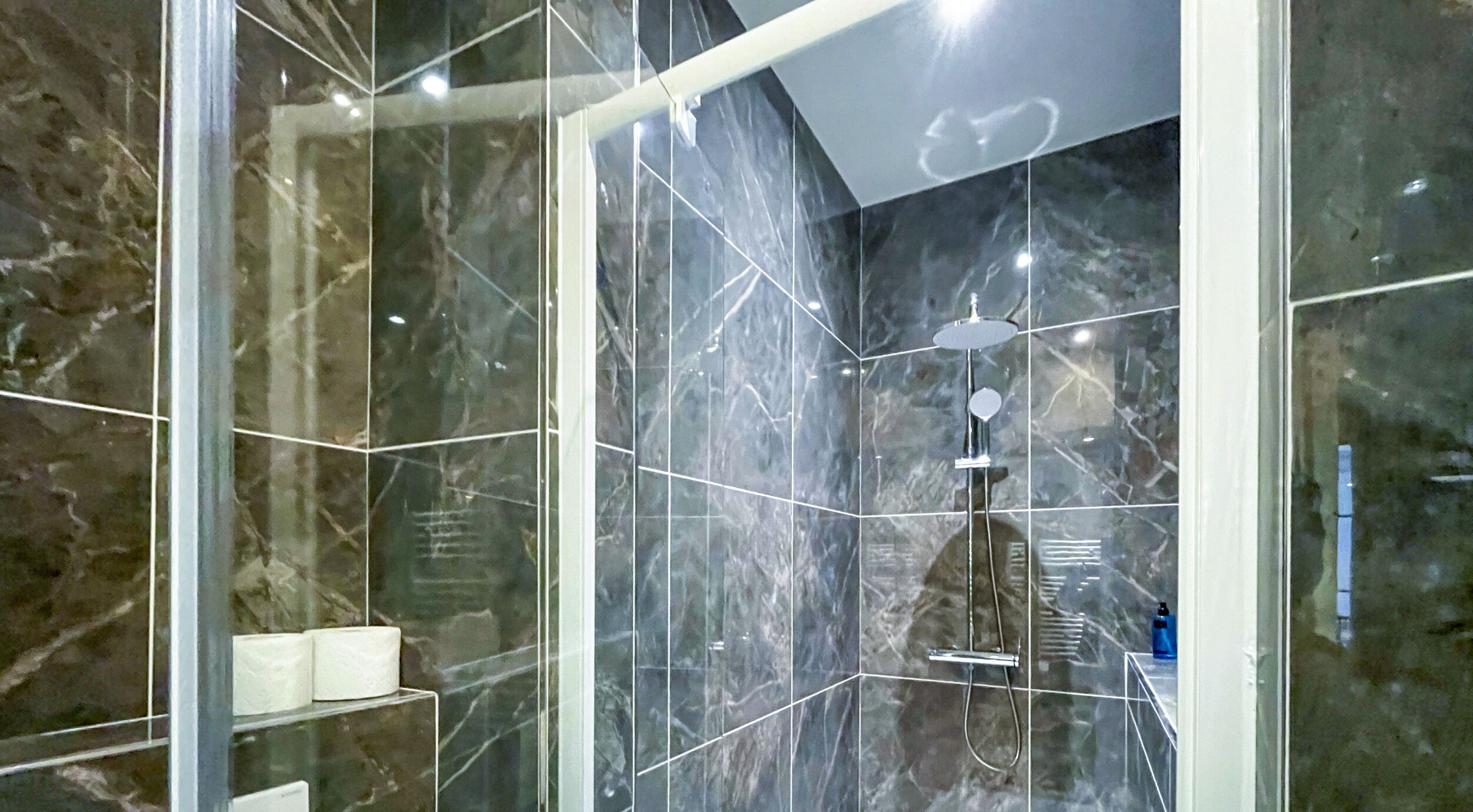 A modern bathroom with a glass-enclosed shower, marble walls, and a visible toilet paper roll.