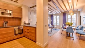 A modern kitchen with wooden cabinets and stainless steel appliances, a dining area with blue chairs and a table, and a living room with a sofa and a window with curtains.