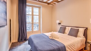 A bedroom with a bed, a window with curtains, a view of buildings, and a wooden ceiling.