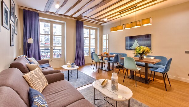 A modern living room with a sofa, dining table, and chairs. There are windows with curtains, a chandelier, and artwork on the wall.