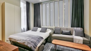A modern hotel room with a bed, a sofa, a wooden side table, and a large window with curtains.