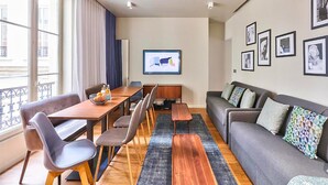 A modern living room with a sofa, a dining table with chairs, a coffee table, and a large window with curtains. - UrbanFlat 79 - Beautiful Large Apartment in Paris Le Marais (París)