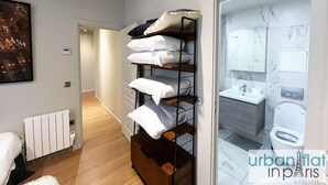 Un dormitorio con suelo de madera, una cama, un radiador, una estantería con almohadas y un baño con inodoro y lavabo.