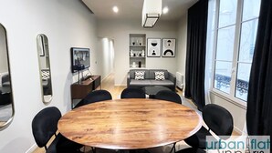 Una sala de estar moderna con una mesa redonda de madera, sillas negras, un sofá gris, un televisor y un ventanal con cortinas.