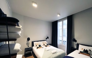 A hotel room with a bed, a bedside table, a window with curtains, and a wall-mounted shelf with towels. - Urban Flat 110 - Luxury 3 Bedroom near Galeries Lafayette (París)