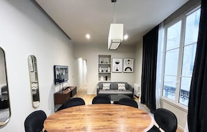 A modern living room with a round wooden table, black chairs, a grey sofa, a TV, and a large window with curtains. - Urban Flat 110 - Luxury 3 Bedroom near Galeries Lafayette (París)