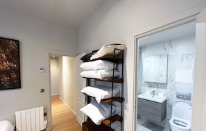 A bedroom with a wooden floor, a bed, a radiator, a shelf with pillows, and a bathroom with a toilet and sink. - Urban Flat 110 - Luxury 3 Bedroom near Galeries Lafayette (París)