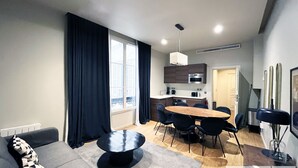 Una sala de estar moderna con sofá, una mesa de comedor con sillas, una zona de cocina con armarios y electrodomésticos, y un ventanal con cortinas.