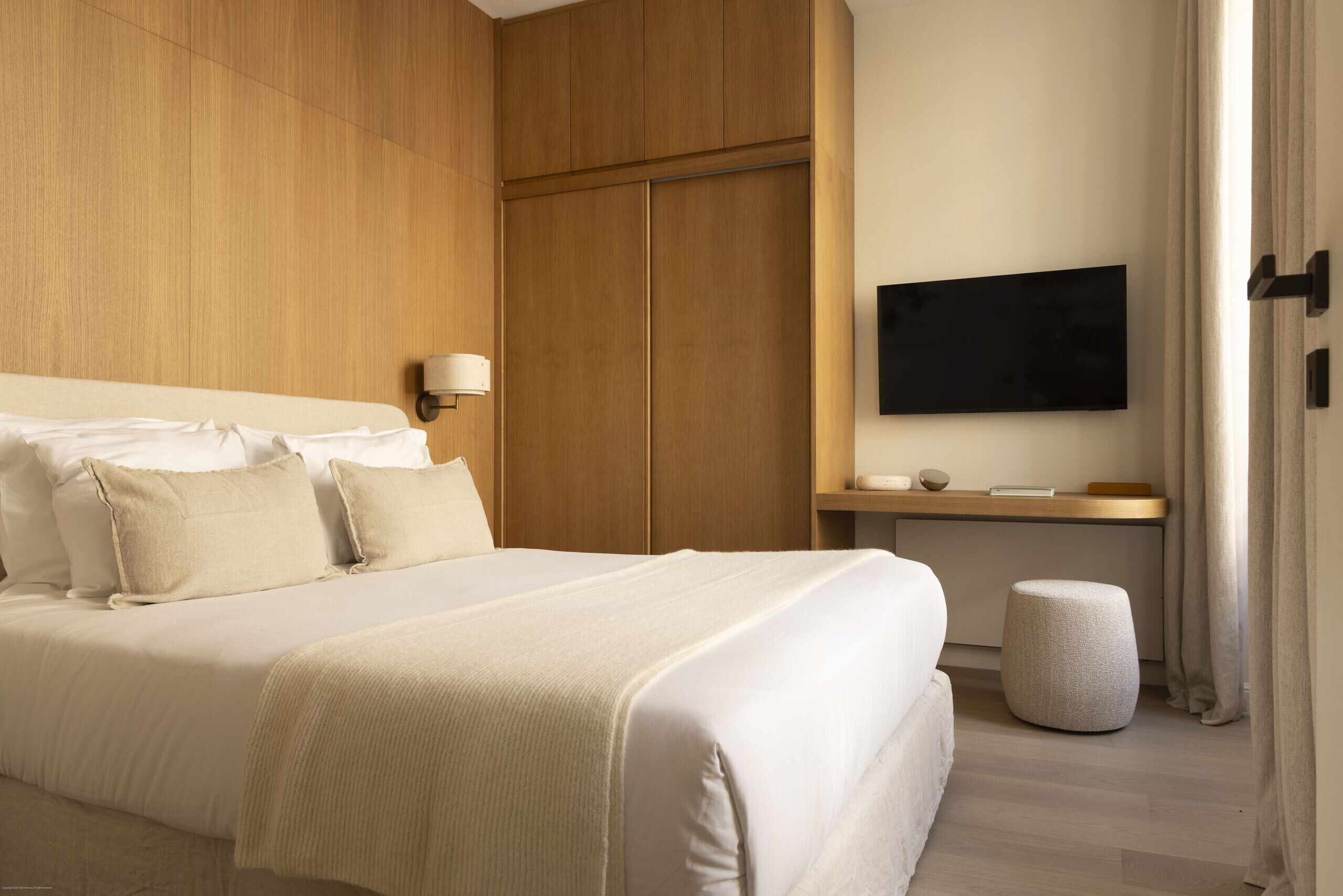 Une chambre d’hôtel avec un grand lit, un téléviseur fixé au mur, une armoire en bois et un petit pouf.
