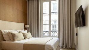 A hotel room with a bed, curtains, a TV mounted on the wall, and a window with a view of buildings.