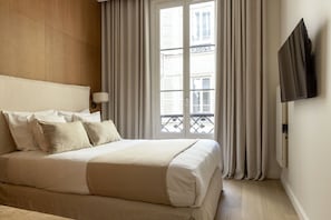 A hotel room with a bed, curtains, a TV mounted on the wall, and a window with a view of buildings.