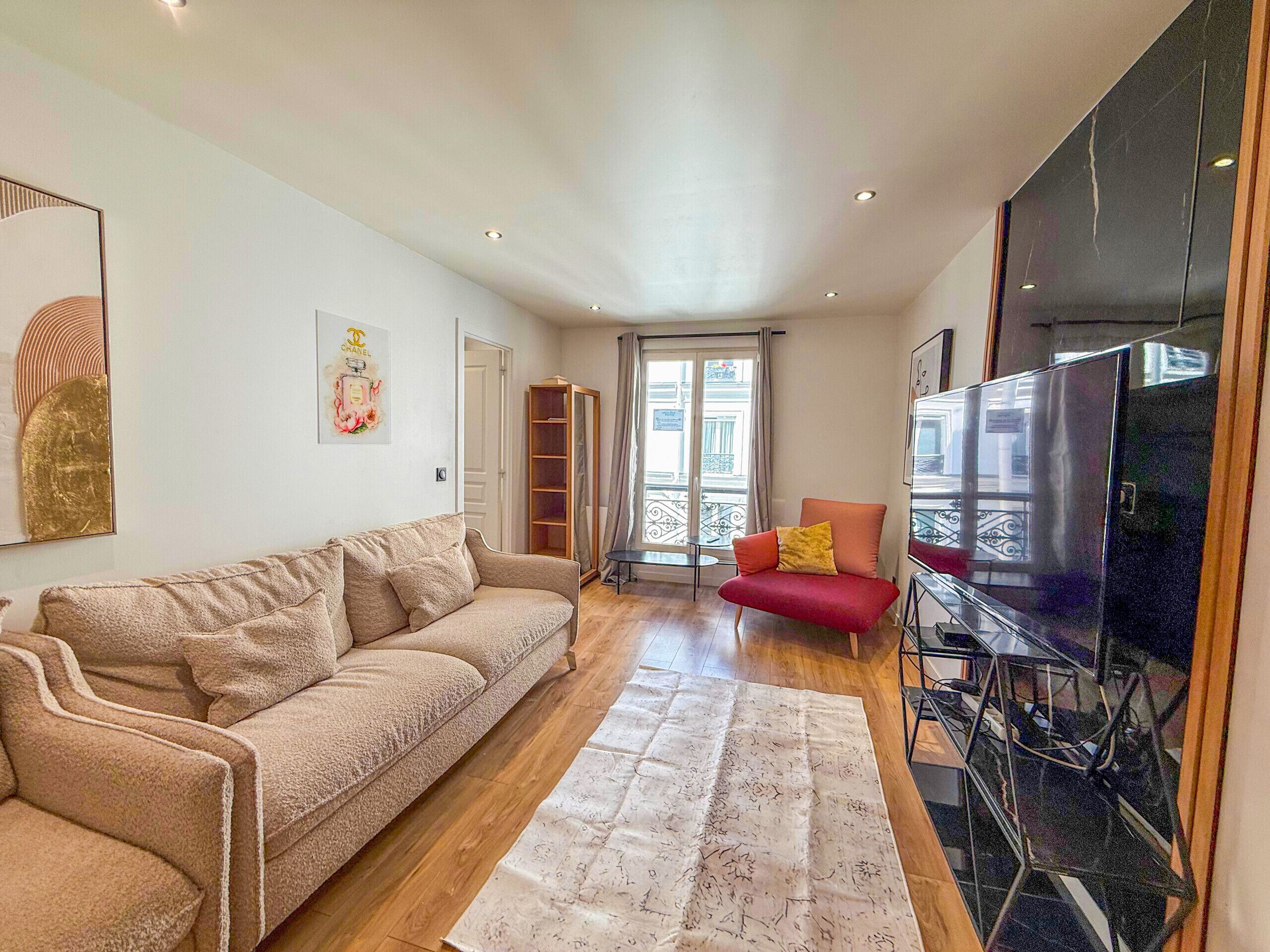 Ein Wohnzimmer mit einem beigen Sofa, einem roten Sessel, einem großen Fernseher und einem Fenster mit Blick nach draußen.