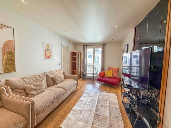 A living room with a beige sofa, a red chair, a large TV, and a window with a view of the outside.