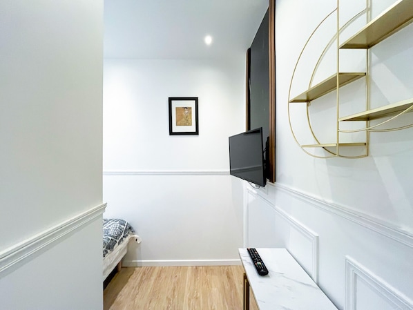A small, modern room with a bed, a mounted TV, a desk with a remote, and a framed picture on the wall.