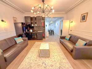 Ein Wohnzimmer mit zwei Sofas, einem Couchtisch und einer Chandelier. Im Hintergrund ist eine Küche mit modernen Geräten und ein Essbereich erkennbar.