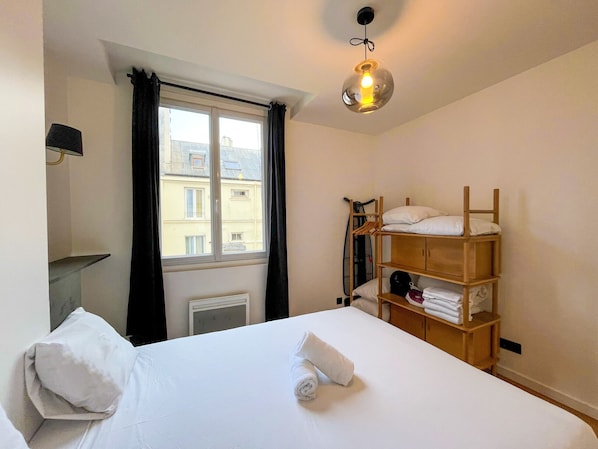 A hotel room with a bed, a wooden shelf with folded towels, a radiator, and a window with curtains.