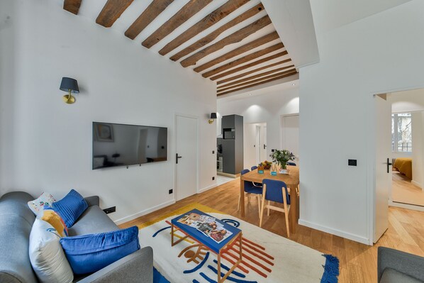 A modern living room with a grey sofa, blue cushions, a coffee table with magazines, a TV mounted on the wall, and a wooden ceiling.