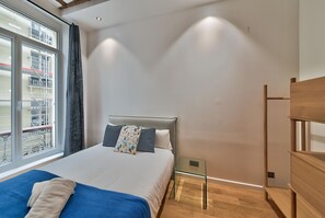 A bedroom with a bed, a glass side table, a wooden dresser, and a window with a view of buildings.