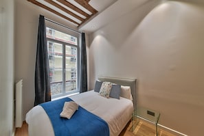 A bedroom with a bed, a glass side table, a window with curtains, and a view of buildings outside.