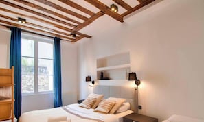 A bedroom with a wooden ceiling, a bed with white bedding, a nightstand, a shelf with items, and a window with curtains.