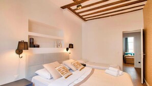 Un dormitorio con cama, mesitas de noche, cabecero, estante y una puerta que da a otra habitación.