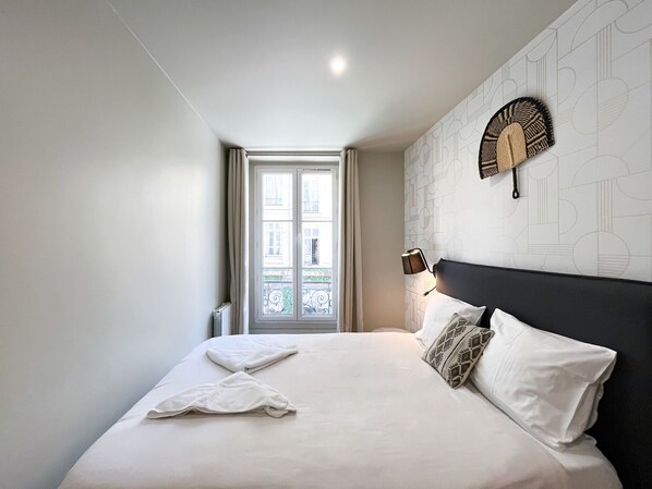 A bedroom with a large bed, a window with curtains, a wall-mounted lamp, and a patterned wall.