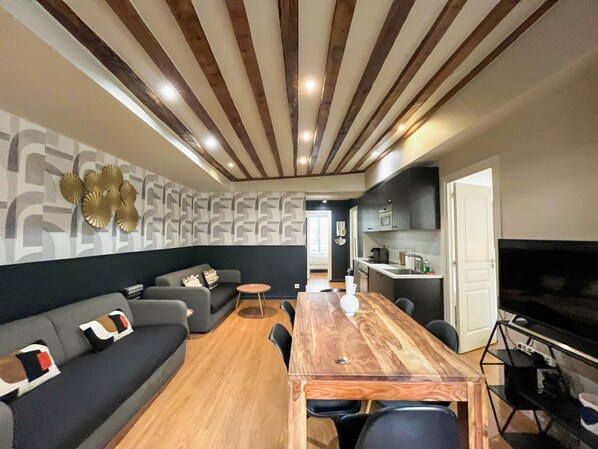 A modern living room with a wooden dining table, grey sofa, and a kitchen area with dark cabinets and a black TV.