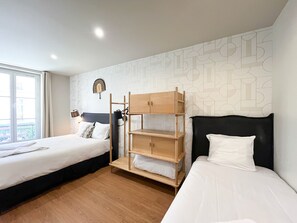 A hotel room with two beds, a wooden bunk bed, a patterned wall, and a window with a view.
