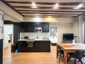 A modern kitchen with dark cabinetry, a microwave, and a built-in oven. There's a dining area with a wooden table and chairs, and a TV in the background.