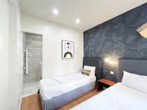 Quarto de hotel com duas camas de solteiro, uma pequena mesa de cabeceira, uma luminária fixada na parede e uma porta para o banheiro.