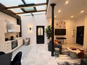 A modern interior with a kitchen, dining area, and living space, featuring a skylight, a large plant, and a flat-screen TV.