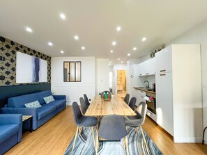 A modern living room with a blue sofa, a wooden dining table with chairs, and a kitchen area with white cabinets and a refrigerator.