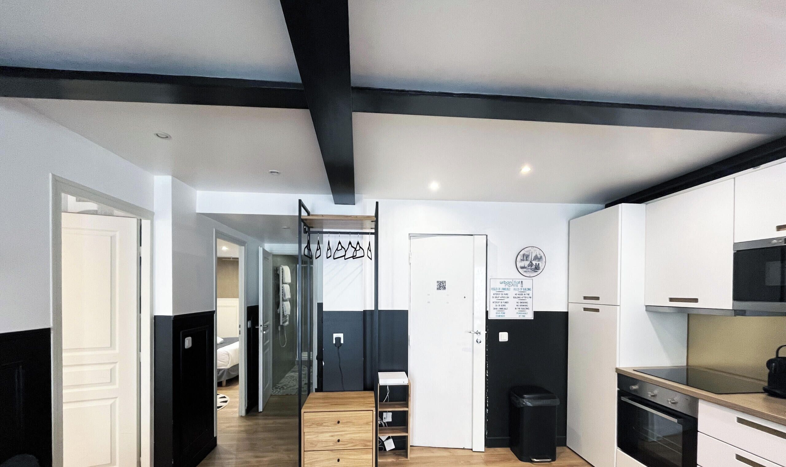 A modern apartment with a kitchen, a dining area, and a living room. There is a wooden floor, white walls, and black accents. A door leads to a bathroom.