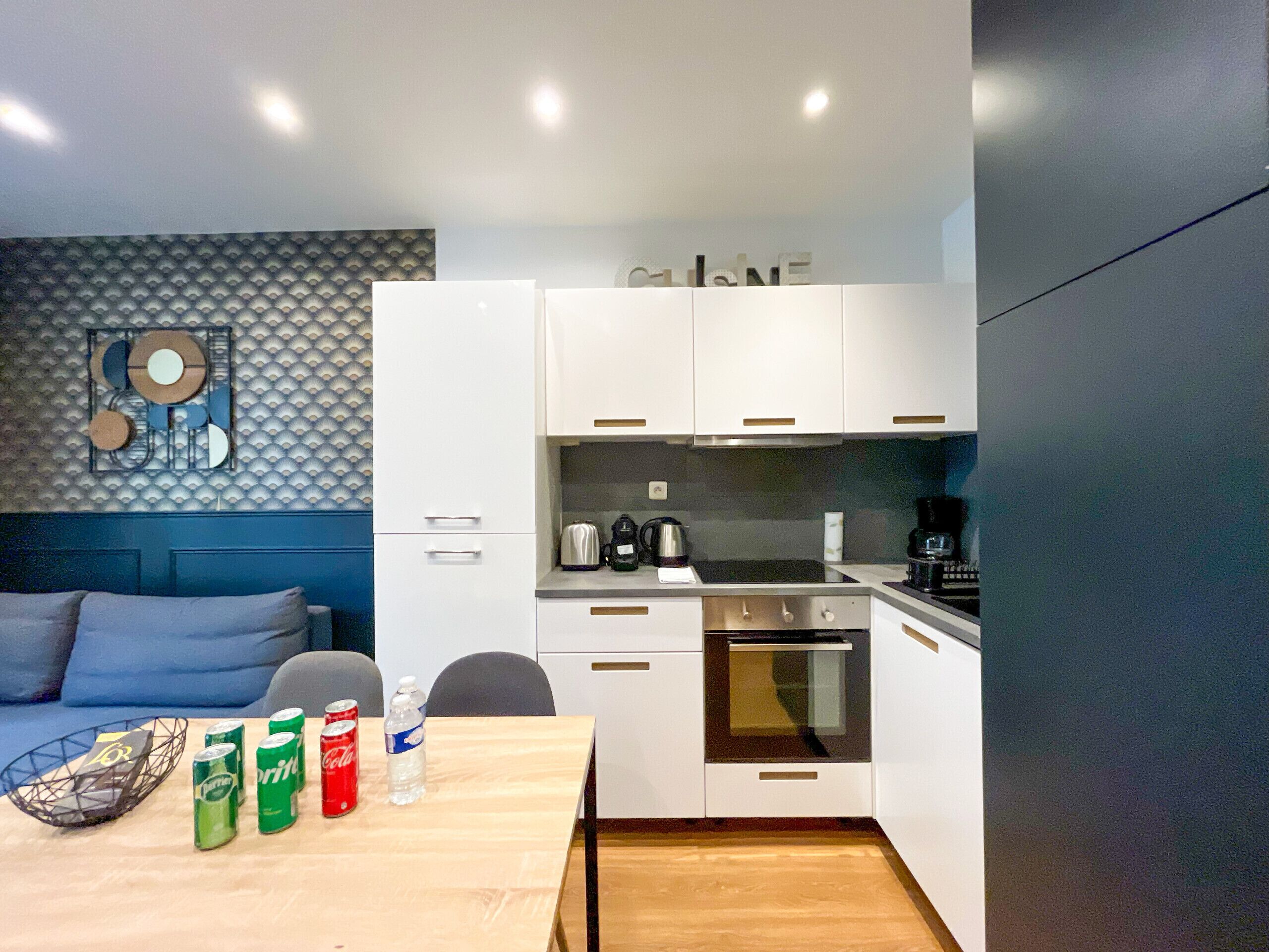 A modern kitchen with white cabinets, a stove, and a coffee maker. There is a dining table with soda cans and a water bottle.