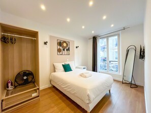 A bedroom with a bed, a fan, a window with a view, and a standing mirror.