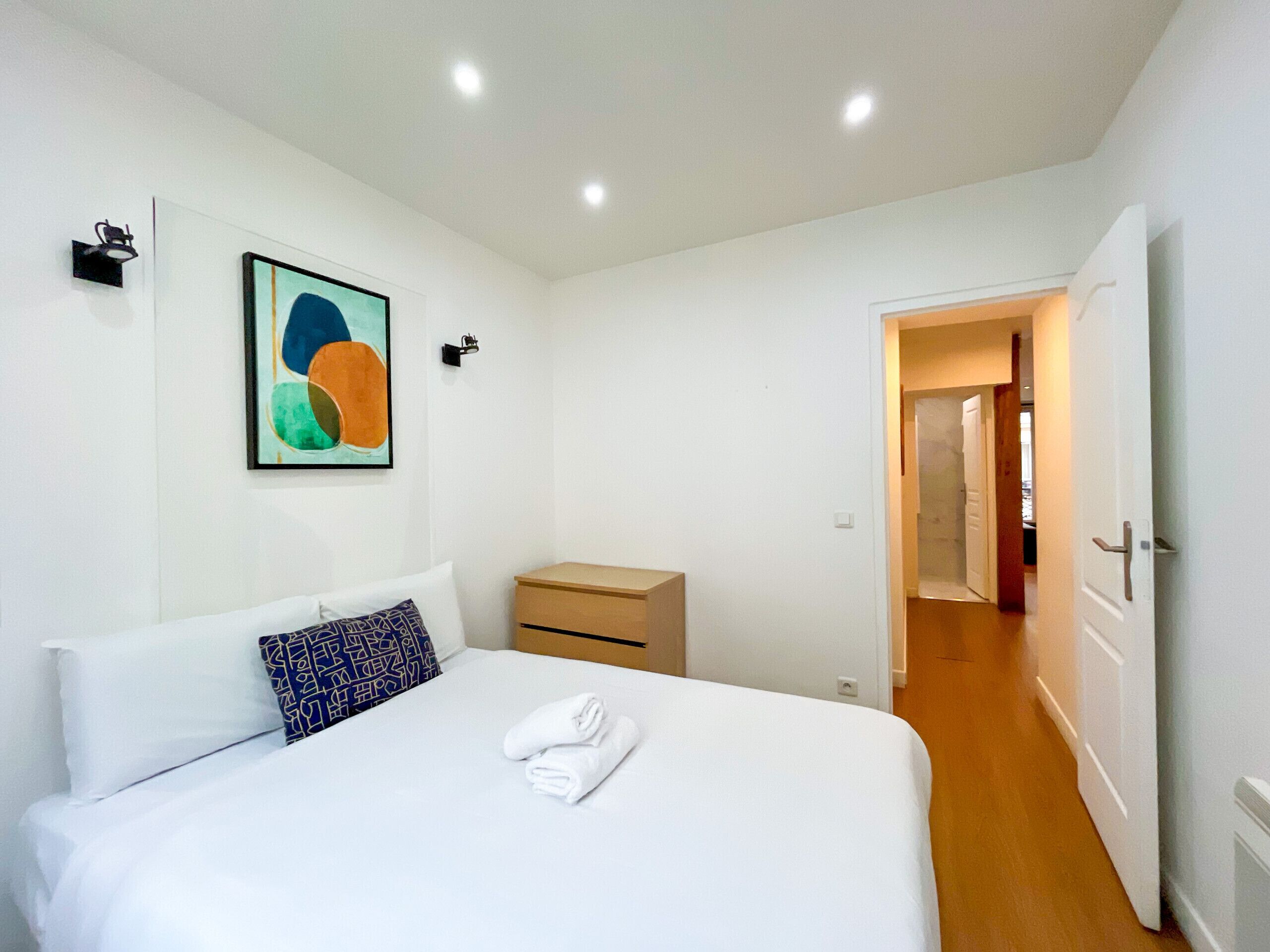 Una habitación de hotel con una cama, una mesita de noche, un cuadro en la pared y una puerta abierta que da a otra habitación.