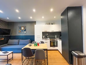 A modern living room with a sofa, dining table, and chairs, a kitchen with white cabinets and appliances, and a black cabinet.