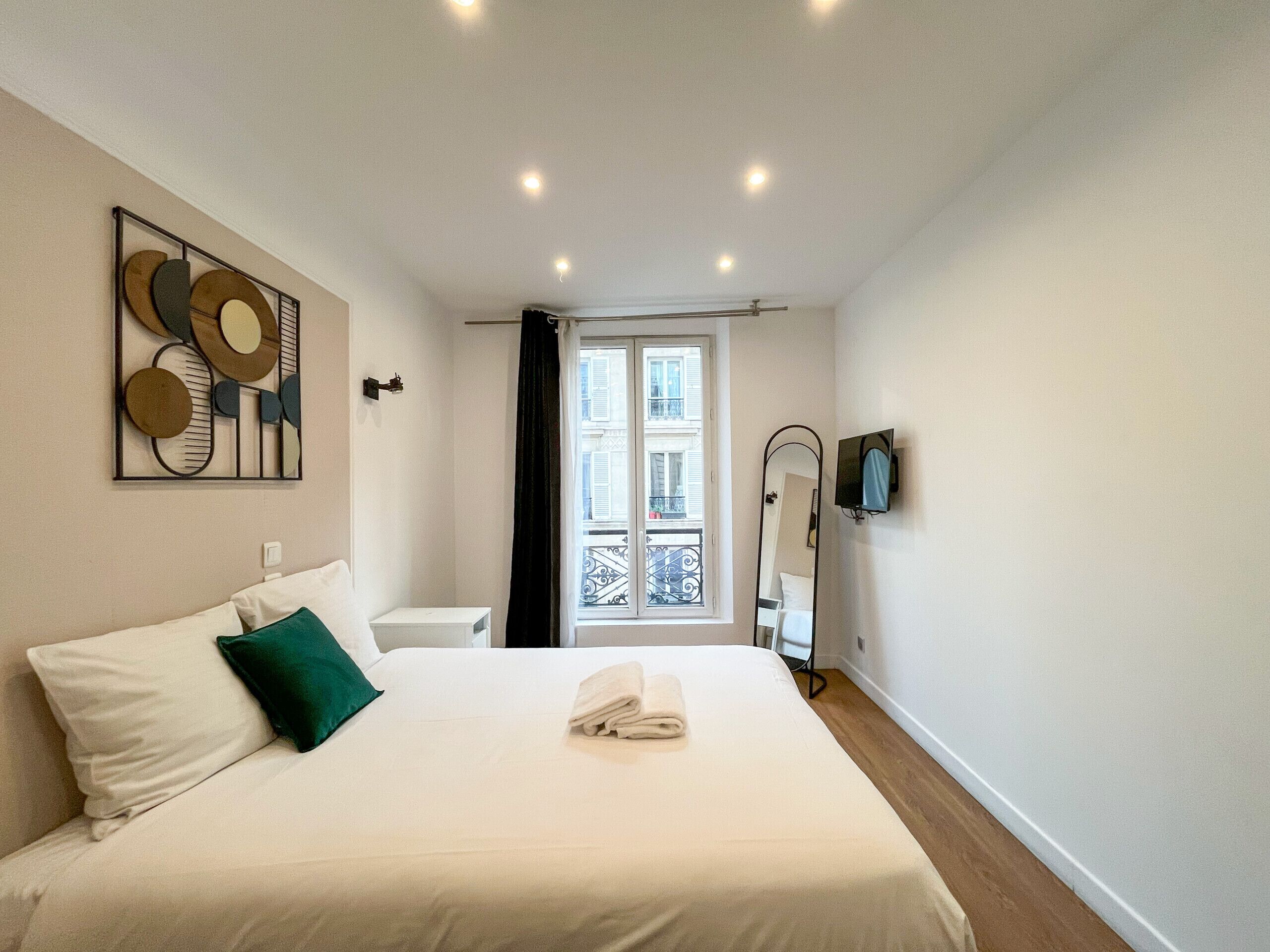 Una habitación de hotel con cama, televisor de pared, ventana con vistas a edificios y una puerta.