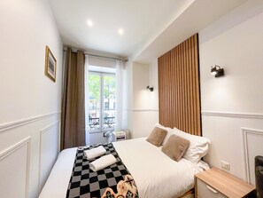 A hotel room with a bed, a wooden panel wall, a window with curtains, and a small bedside table.