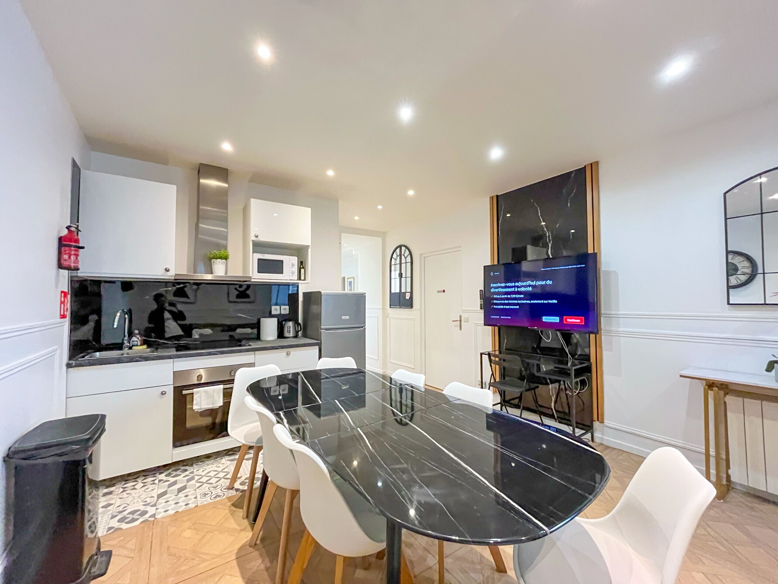 Een moderne keuken met witte kastjes, een zwart aanrecht en een roestvrijstalen koelkast. Er staat een eettafel met witte stoelen en een flatscreen televisie is aan de muur bevestigd.