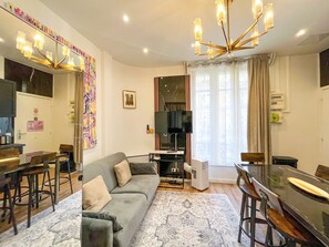 A modern living room with a grey sofa, a dining table with chairs, a TV, and a chandelier.