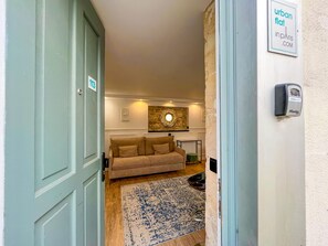 A hallway leading to a room with a beige sofa, a patterned rug, and a wall-mounted light fixture.