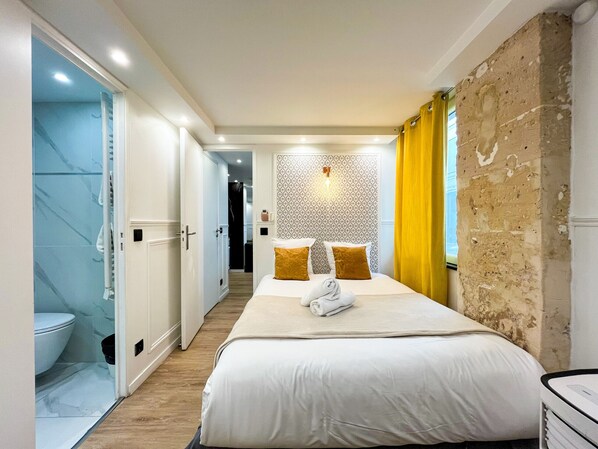 A hotel room with a bed, a bathroom with a toilet, a shower, and a hallway leading to another room.