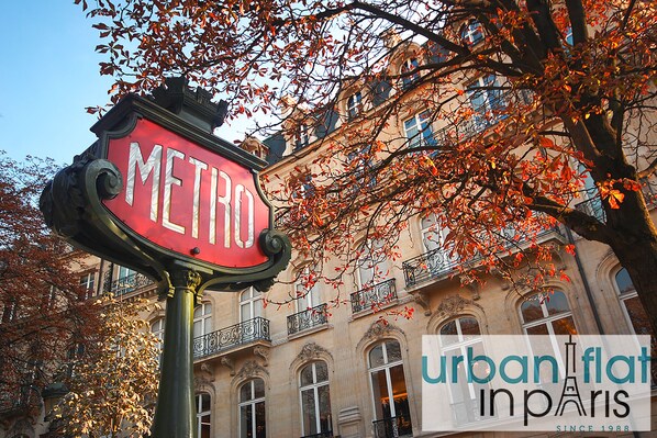 Un cartel con la palabra "METRO" en un poste, con un edificio y hojas otoñales al fondo.