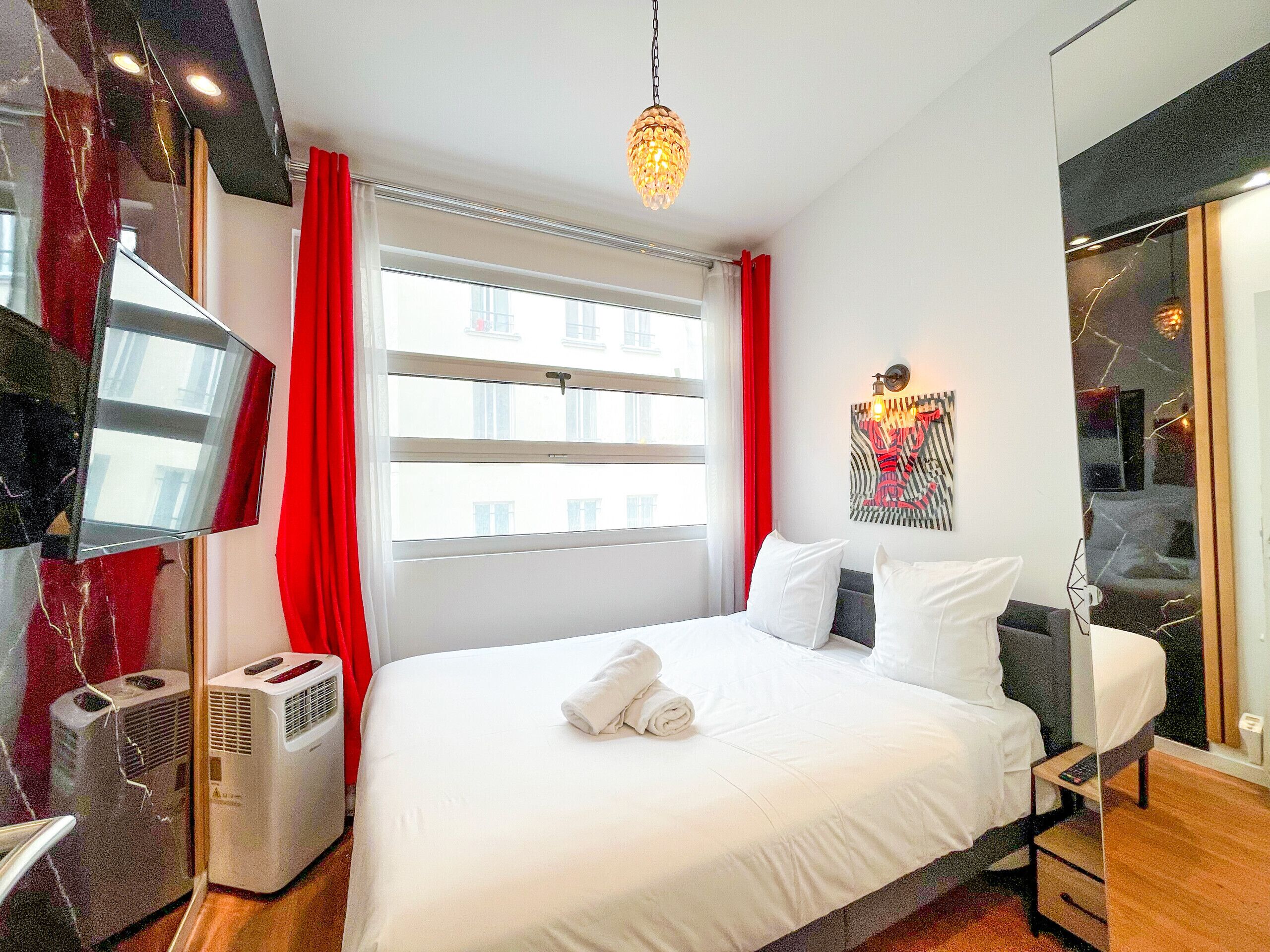 A hotel room with a large bed, a wall-mounted air conditioner, a window with red curtains, and a modern light fixture.