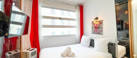 A hotel room with a large bed, a wall-mounted air conditioner, a window with red curtains, and a modern light fixture.