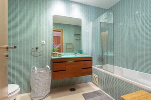 Une salle de bain avec un mur recouvert de carreaux verts, un meuble-lavabo en bois, une baignoire blanche et un sac à ordures à rayures.